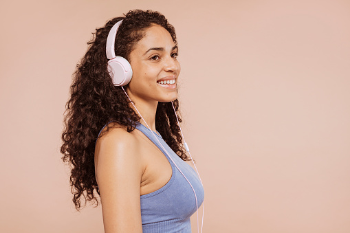 Side view of young multiracial cheerful woman, dressed in gym outfit, listens music with modern headphones, poses at studio isolated over beige background. Fitness and music concept.