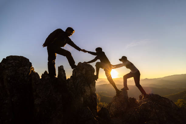 silhouette du travail d’équipe de trois randonneurs s’entraidant au sommet de l’équipe d’alpinisme. travail d’équipe amitié randonnée s’entraider faire confiance assistance silhouette en montagne, lever du soleil. - rescue mountain horizontal three people photos et images de collection