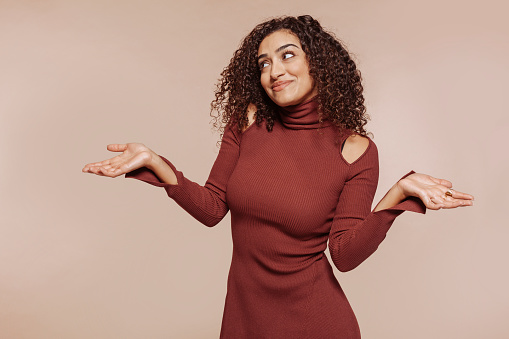 Young beautiful arabic smiling woman with curly hair, wears garnet dress at studio isolated over beige background. Clueless, confused, having doubts expression with arms and hands raised. Doubt concept.