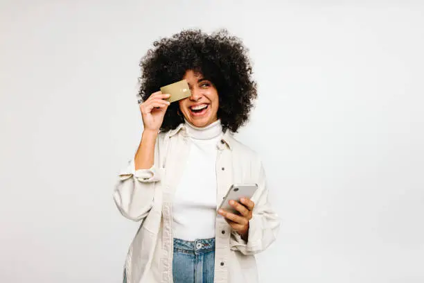 Cheerful young woman smiling happily while holding a credit card and a smartphone. Gorgeous woman with curly hair enjoying some online shopping while standing against a white background.