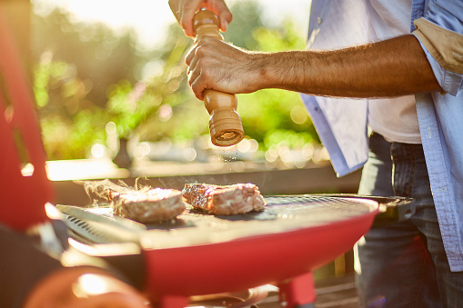 Close up on man's hand seasoning meat on the gas grill on barbecue grill outdoor in the backyard, grilled roasted steak meat, summer family picnic, food on the nature.