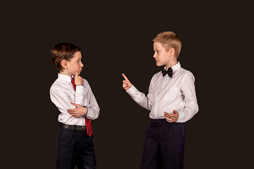 Two confident serious boys friends in white shirt talking isolated on black background, looking at each other speaking. Children debating argument. Business discussion concept. Copy text space for ad