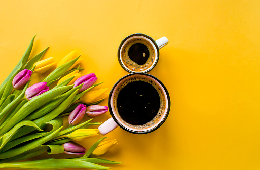 Banner.Two cups of hot, morning coffee in the shape of the number 8 and a bouquet of yellow-lilac tulips on a bright yellow background. View from above.Copy space for text. The concept of holidays.