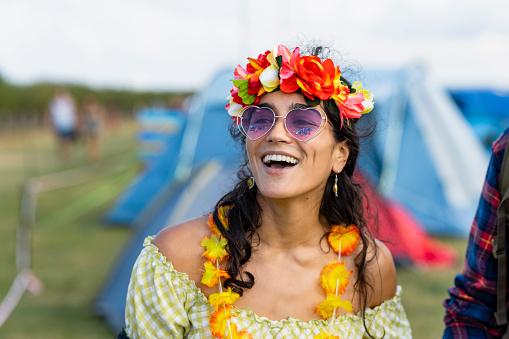 A young woman at a festival in Lindisfarne, North East England. She is wearing a flower headband and garland and heart sunglasses while laughing and looking away from the camera slightly.