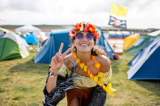 A young woman at a festival in Lindisfarne, North East England. She is wearing a flower headband, flower garland and heart sunglasses and doing the peace sign while looking and smiling at the camera.
