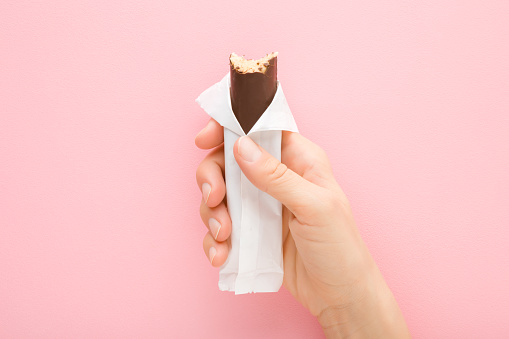 Young adult woman hand holding bitten dark brown chocolate bar with marzipan on light pink table background. Pastel color. Closeup. Sweet snack in opened white pack. Top down view.