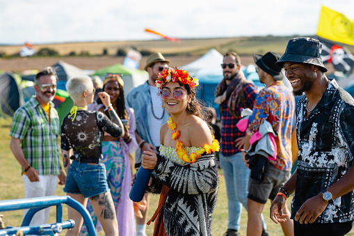 Festival goers at a festival in Lindisfarne, North East England. The main focus is a young woman wearing festival gear and carrying a reusable water bottle while walking through the festival field and smiling.