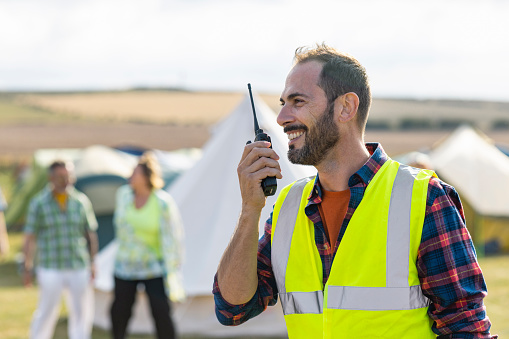 A volunteer wearing a reflective vest and working at a festival in Lindisfarne, North East England. He is talking to someone using a walkie talkie while smiling.