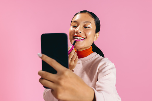 Female using a smartphone as a mirror to apply lipstick in a studio. Gen Z woman doing the two toned lip trend to create a bold and vibrant makeup look on her face.