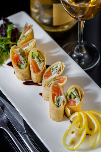 Pancake rolls with salmon, Philadelphia cheese and dill. The rolls are on a white, rectangular plate, next to it is a mix of salad and lemon slices. The plate stands on a wooden background. there are cutlery next to it. On the other side is a glass of white wine and a bottle.