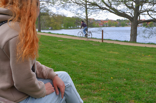 Red-haired woman sits on a park bench near the lake. Cropped photo. Rear View Of Woman Sitting On Bench Against Lake