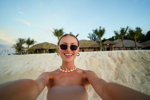 Young beautiful blonde woman in sunglasses smiling to the camera while taking selfie on the sandy tropical beach. Girl enjoying summer vacation concept. Travel. Tourist.