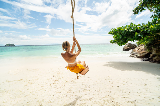 Happy woman on the tropical beach having fun on swing, sea shore during summer vacations. Copy space. Traveler. Tourist. Wanderlust. Real people lifestyle.