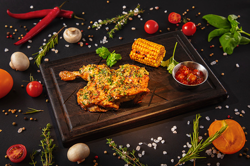 Fried half of chicken with sauce, parsley and corn on a wooden board with mushrooms, peas, red chili peppers, salt, tomatoes and basil on a black background