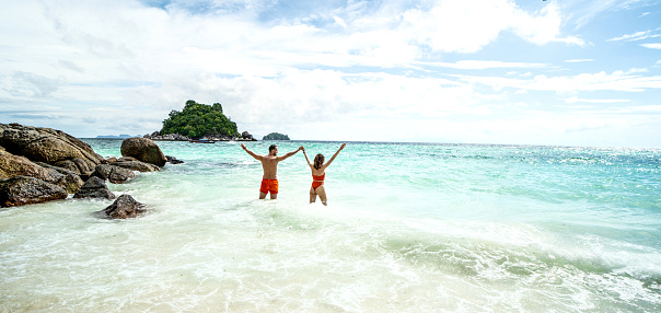 Happy couple having fun on the tropical beach, standing in the water, relaxing, sunbathing. Back view. Copy space. Travelers. Tourists. Nature. Island vibes.
