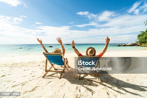 istock Two happy people having fun on the beach, sitting on blue sunbed with hands raised up, spending leisure time together. Summer holidays concept. 1458771594