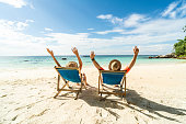 Two happy people having fun on the beach, sitting on blue sunbed with hands raised up, spending leisure time together. Summer holidays concept.