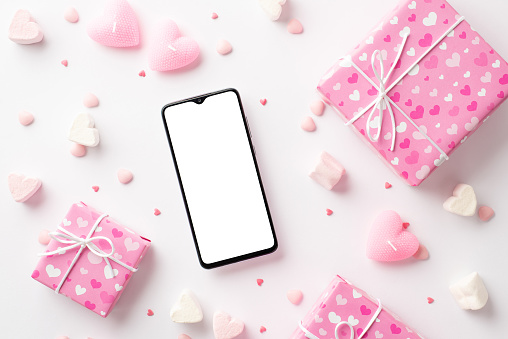 Valentine's Day concept. Top view photo of smartphone pink gift boxes heart shaped marshmallow candles and sprinkles on isolated white background with blank space