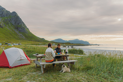Happy heterosexual couple contemplating an outdoor dinner by the mountain beach campsite during summer sunset on Lofoten, Norway