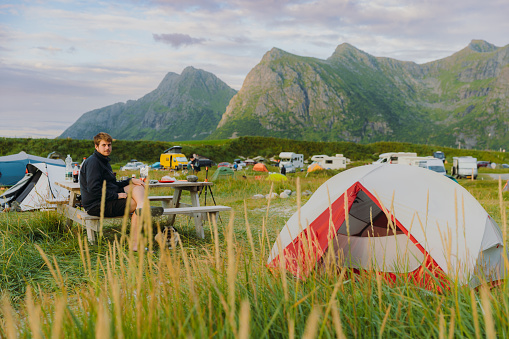 A male traveler contemplating an outdoor dinner by the mountain beach campsite during summer sunset on Lofoten, Norway