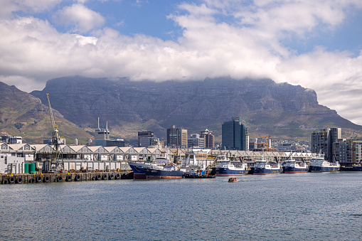 Cape Town, South Africa - December 14th 2022: Row of fishing boats in the harbor with a view to the cloud covered Table Mountain in the back