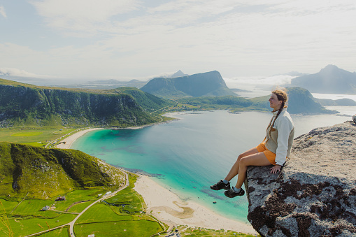 Happy female sitting at the edge of the mountain, admiring a view of the turquoise beach and dramatic mountain peaks during summertime on Lofoten, Northern Norway