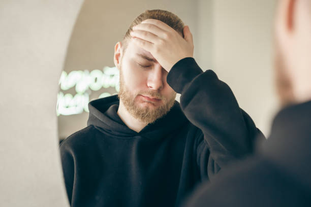 Sad young man with a beard in front of a mirror, mental health concept. Sad, tired young man with a beard in front of a mirror, covers his face with his hand, mental health concept. psyche photography stock pictures, royalty-free photos & images