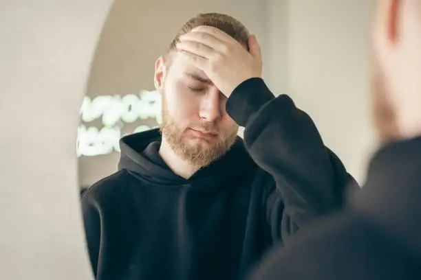 Sad, tired young man with a beard in front of a mirror, covers his face with his hand, mental health concept.