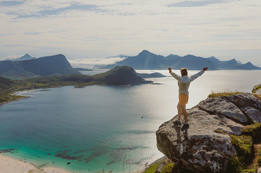 Happy female with arms raised staying at the edge of the mountain, admiring a view of the turquoise beach and dramatic mountain peaks during summertime on Lofoten, Northern Norway