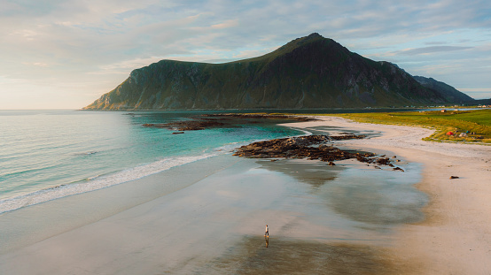 High-angle drone photo of a female traveler admiring the bright summer sunset walking at the sand beach with view of the ocean and the mountain peaks in Northern Norway