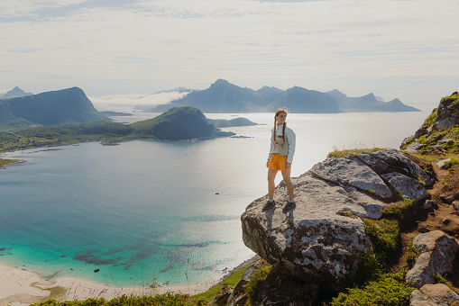 Happy female staying at the edge of the mountain, admiring a view of the turquoise beach and dramatic mountain peaks during summertime on Lofoten, Northern Norway