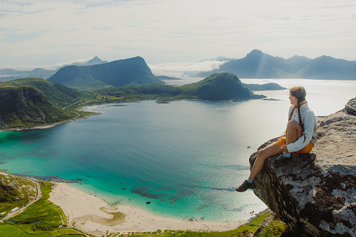 Happy female sitting at the edge of the mountain, admiring a view of the turquoise beach and dramatic mountain peaks during summertime on Lofoten, Northern Norway