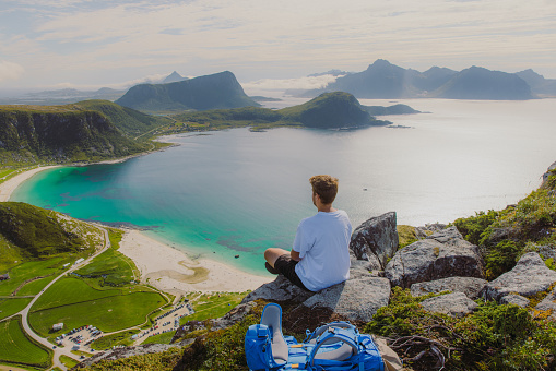 Scenic view of a male sitting at the edge of the mountain, admiring a view of the turquoise beach and dramatic mountain peaks during the summertime on Lofoten, Northern Norway
