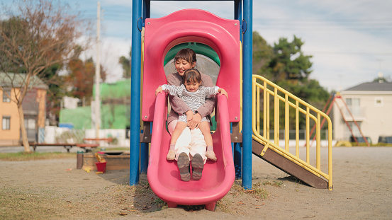 A small girl is playing with her mother in a public park and sliding on a slide.