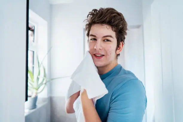 Portrait of smiling teenage boy with acne problem who takes care his face skin at home. He looking in the mirror and wipes face with towel in bathroom. Teenager skin care every day treatment process