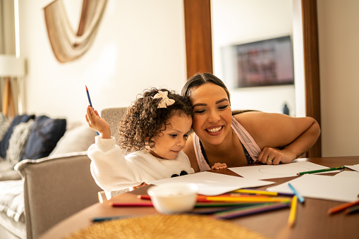 Mother hugging her daughter while she drawing on paper at home