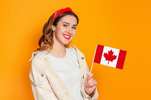 Young girl student smiling and holding a small canada flag and looking away isolated over orange background, Canada day, holiday, confederation anniversary, copy space