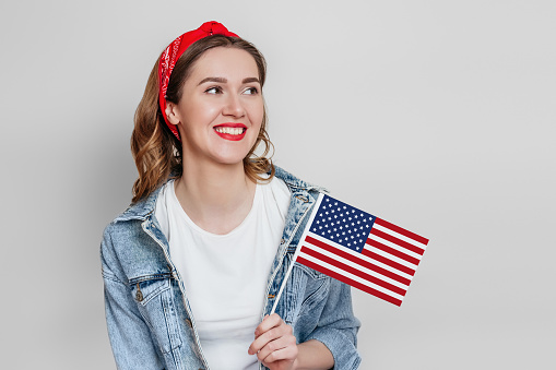 Happy young woman with red lipstick holds a small american flag and smiles isolated over grey background, girl holding USA flag, 4th of july independence day, copy space