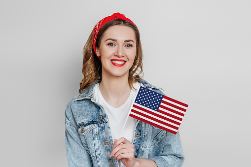 Happy young girl student with red lipstick holds a small american flag and smiles isolated over grey background, girl holding USA flag, 4th of july independence day, copy space