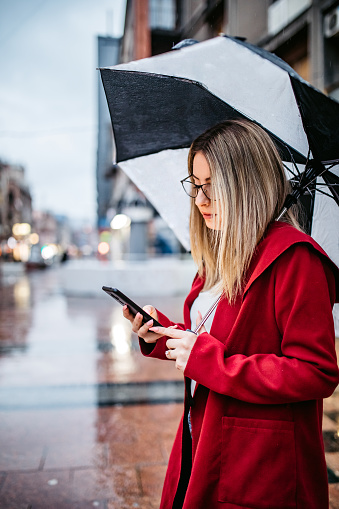 Beautiful young woman standing on the street, holding an umbrella and using her smart phone as the rain pours.