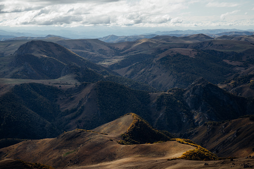 Photo of a scenic view of valley and hills under sunlight and clouds shadows from the plateau Bermamyt. There are trees in autumn colors on the top of the hills and the gorges. Plateau Bermamyt it's a popular touristic place in Karachay-Cherkessia in Russia. Big Bermamyt is located at an altitude of 2591 meters there you can enjoy panoramic views of the Caucasian Ridge and Elbrus