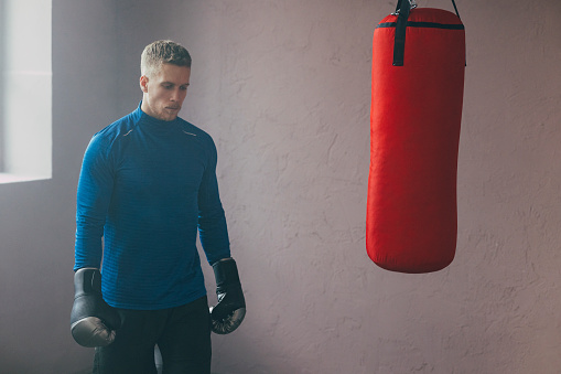 A handsome Caucasian athlete in sports clothes standing near punching bag during his daily boxing workout in the gym.