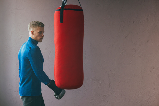A side view of a handsome Caucasian athlete in sports clothes standing near punching bag during his daily boxing workout in the gym.