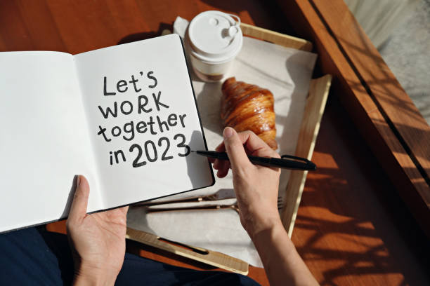 Closeup woman's hands writing down inspirational quote 'let's work together in 2023' on a note pad with coffee and croissant on background.