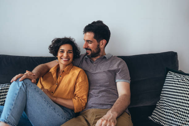 A Happy Beautiful Couple Looking At Camera While Sitting On The Cozy Sofa And Enjoying Spending Their Leisure Time Together A portrait of a pretty Latin-American woman and her smiling Caucasian boyfriend relaxing at home. adult beautiful caucasian love stock pictures, royalty-free photos & images