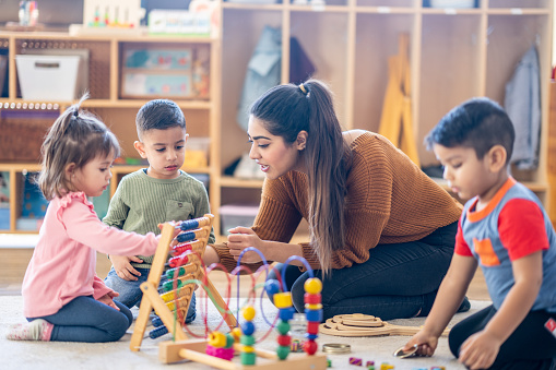 A female Kindergarten teacher of Middle Eastern decent, sits on the floor with students as they play with various toys and engage in different activities.  They are each dressed casually as they learn through their play.