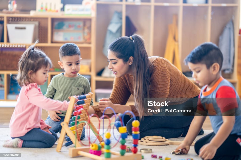 Learning Through Play A female Kindergarten teacher of Middle Eastern decent, sits on the floor with students as they play with various toys and engage in different activities.  They are each dressed casually as they learn through their play. Child Care Stock Photo