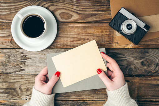 An envelope and a blank sheet of paper in female hands on a wooden background with a retro camera and a cup of coffee, top view.