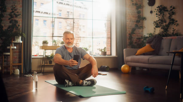 starke athletische passform mann mittleren alters mit smartphone zwischen den trainingseinheiten während der morgengymnastik zu hause in sonniger wohnung. gesunder lebensstil, fitness, erholung, wohlbefinden und ruhestand. - senior adult healthy lifestyle athleticism lifestyles stock-fotos und bilder