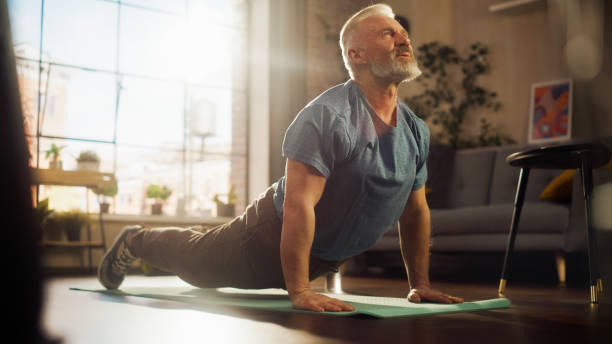 Strong Athletic Fit Senior Man Training on a Yoga Mat, Doing Back Stretching and Core Strengthening Exercises During Morning Workout at Home in Sunny Apartment. Concept of Health and Fitness. fuerte entrenamiento atlético para personas mayores en una esterilla de yoga, haciendo ejercicios de estiramiento de la espalda y fortalecimiento del núcleo durante el entrenamiento matutino en casa en sunny apartment. concepto de salud y fitness.